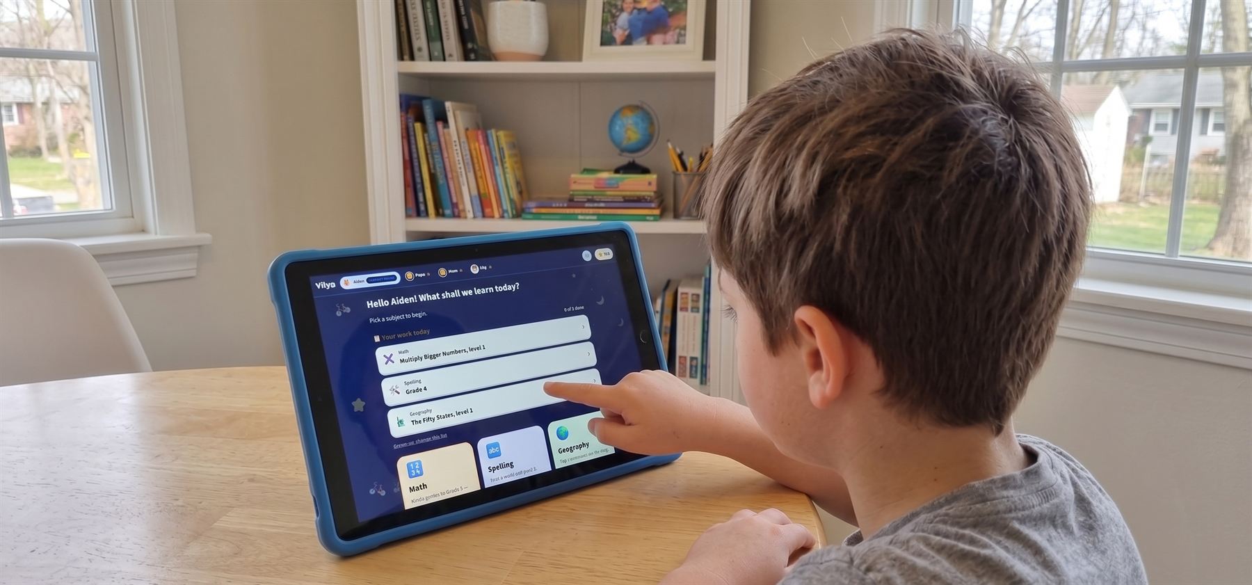 A boy at a kitchen table, reaching out to tap the day's work on a tablet running the app.